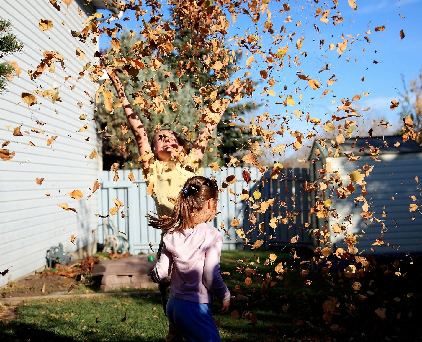 A child laughing while throwing a handful of autumn leaves into the air in a backyard.